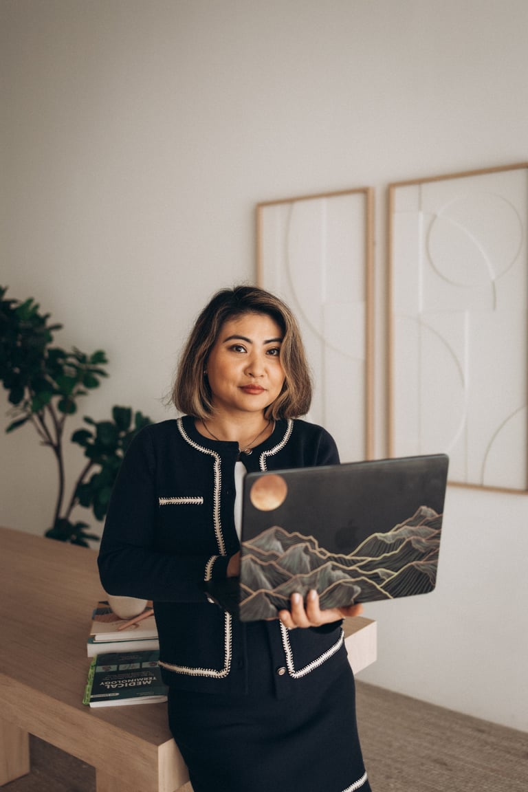 Woman in black cardigan holding laptop with mountain design, sitting at desk with plant and framed art in background